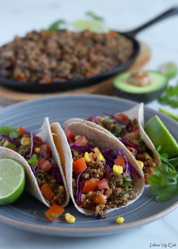 Close up of three folded tacos on a blue-grey plate that are filled with vegan taco meat, tomato, corn, red cabbage and cilantro along with lime slices and cilantro garnishing the plate. Blurred oval cast iron pan on wood board in background with half an avocado to the right.