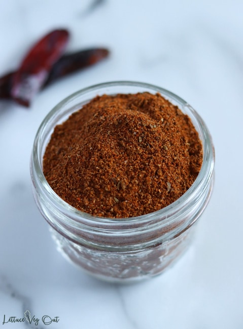 Close up of a heaping glass jar filled with dark red taco seasoning mix. Two dry red chili peppers sit blurred in the back left corner.