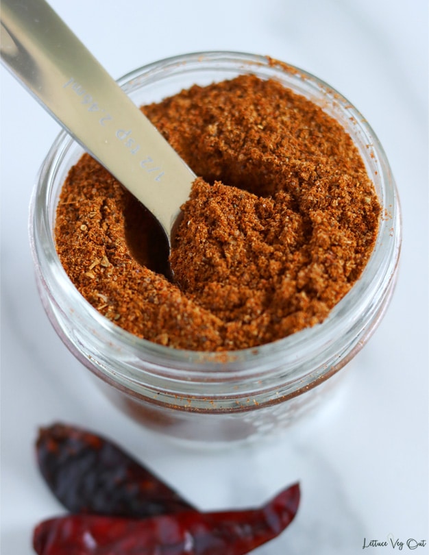 Close up of a small round jar filled with dark red taco seasoning with a metal measuring spoon sitting in the jar. Two dry red chili peppers sit in the bottom left corner of image.