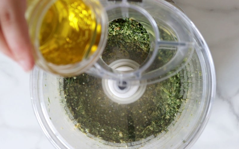 Top view of a food processor that has a partially blended mixture of pesto in it. The lid is on the processor but the pouring hole is open and a hand holding a small jar of oil is about to pour the oil into the food processor.
