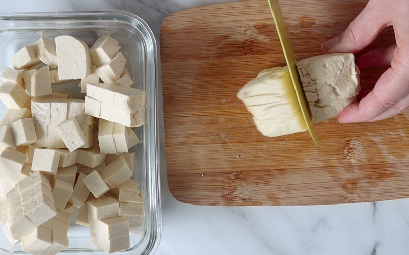 Top view of a small wood cutting board where a block of tofu is being cut with a yellow knife. On the left side of the image is a rectangular glass container filled with tofu cubes.