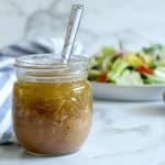 Square cropped image of Straight on view of small glass jar of Italian dressing with spoon in it. White-blue striped towel to the left and wrapped behind the jar with a plate of salad blurred to the back right. White-grey marble background.