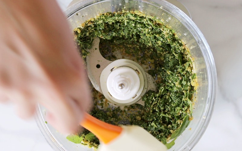 Top view of a food processor with a crumbly looking mixture of basil and nuts in it. A blurry hand is using an orange spatula to scrape the edges of the food processor.