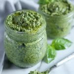 Square cropped image of two jars of pesto sauce, topped with a drizzle of olive oil sit on white-grey marble with a blurred grey towel crumpled behind them. The front jar is slightly larger than the back one. A spoonful of pesto rests in front of the jars.