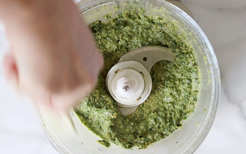 Top view of a food processor filled with blended pesto sauce. A hand is scraping the edges of the food processor (hand is blurry).