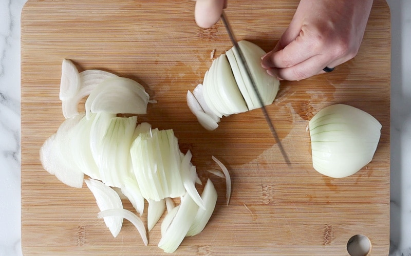 Top view of a cutting board with onion slices and larger onion halves. Hands are slicing an onion (one hand holding a knife and the other holding the onion).