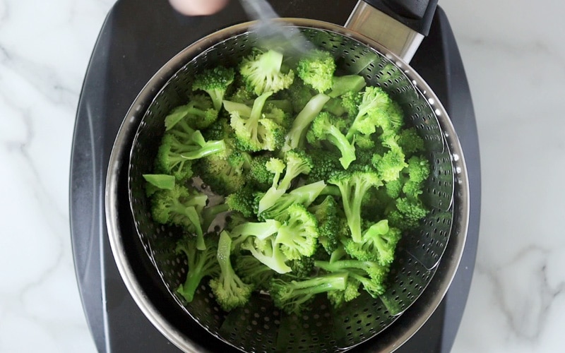 Top view of a pot with a steaming basket inside. The basket is filled with bright green, partially cooked, broccoli florets.