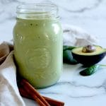 Square cropped image of one glass mason jar of green apple cinnamon smoothie with some of its ingredients around the jar, including two cinnamon sticks, half an avocado and spinach leaves