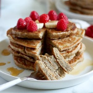 Square cropped image of a close up of a bite of a banana pancake stack on a fork with the rest of the stack behind the fork. Pancakes are topped with raspberries, banana and syrup. Blurred decorations in the back (second stack of pancakes, dish of raspberries, bowl of banana slices).
