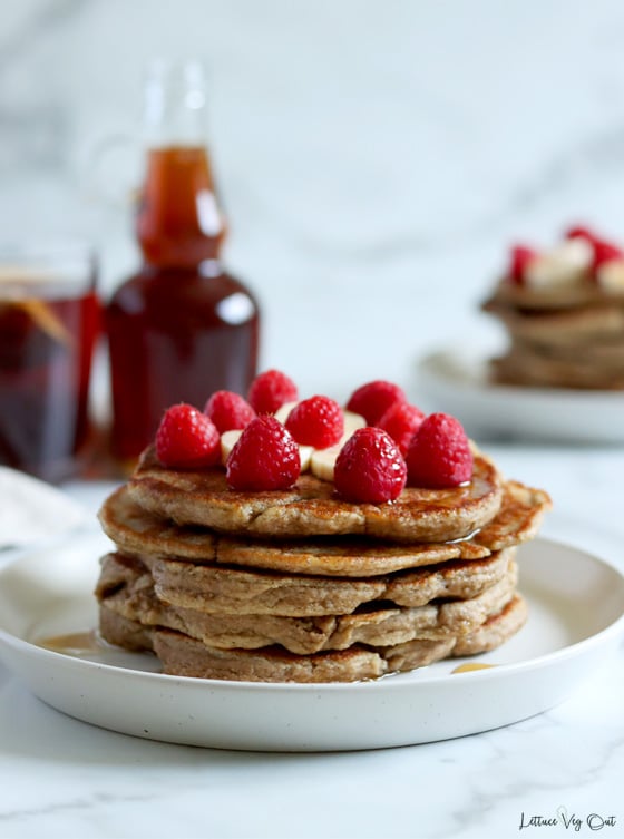 Straight on view of a stack of 5 pancakes topped with raspberries and banana with a blurred second stack of pancakes in the back right. To the back left is a blurred mug of tea and jar of maple syrup.