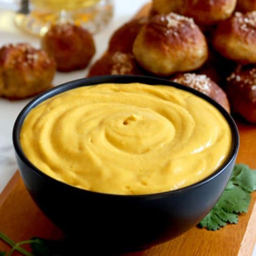 Square cropped image of a black bowl filled with beer cheese dip sits on wood board with pile of pretzel bites stacked on the board behind the bowl. Glass mug of golden beer sits to the back left of the board with two pretzel bites in front of the mug