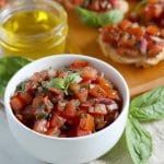 Square cropped image of a small white bowl filled with prepared tomato bruschetta mix sitting in front of a long wood board topped with bruschetta pieces made on baguette with lots of basil leaves for garnish.
