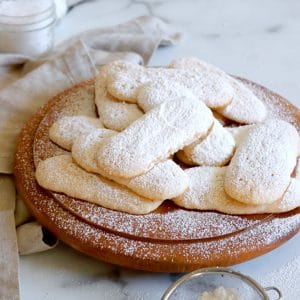 Square cropped image of Pile of ladyfinger cookies, topped with sprinkled icing sugar, on a round wood board with white-grey marble background.