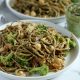 Square cropped image of a close up of a plate of pesto pasta (spaghetti) loaded with broccoli, chickpeas and caramelized onions with a sprinkle of parmesan cheese on top. A second plate of pasta is blurred in the back and a small jar of pesto sauce sits between the plates to the right.