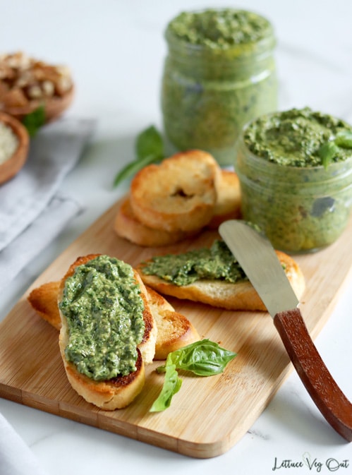 A small wood board is topped with toasted crostini pieces, some of which are topped with a bright green pesto sauce. Two differently sized jars of pesto sit to the back of the board and there is a small butter knife resting on a crostini in the center of the board. Small wood dishes sit blurred in the back left.