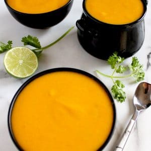 Square cropped image of three small black bowls of creamy pumpkin coconut soup sitting on white-grey marble that is decorated with cilantro sprigs and lime.