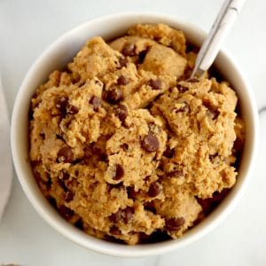 Square cropped image of the Top view of white bowl filled with raw chickpea cookie dough with mini chocolate chips. Marble spoon in cookie dough