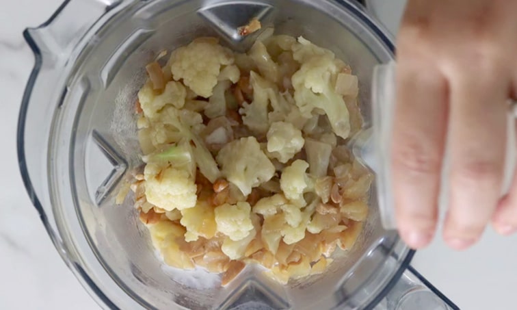 Top view of a blender filled with cooked cauliflower, onion and cashews with a hand pouring some liquid into the blender on the right side of image.