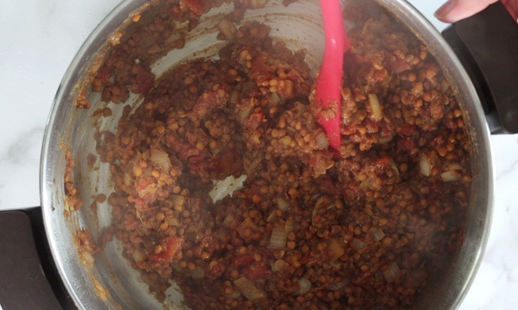 Top view of a large metal pot filled with cooked lentil sloppy joes that are releasing steam (and slightly blurring the image) while a large red spoon is stirring the mixture.