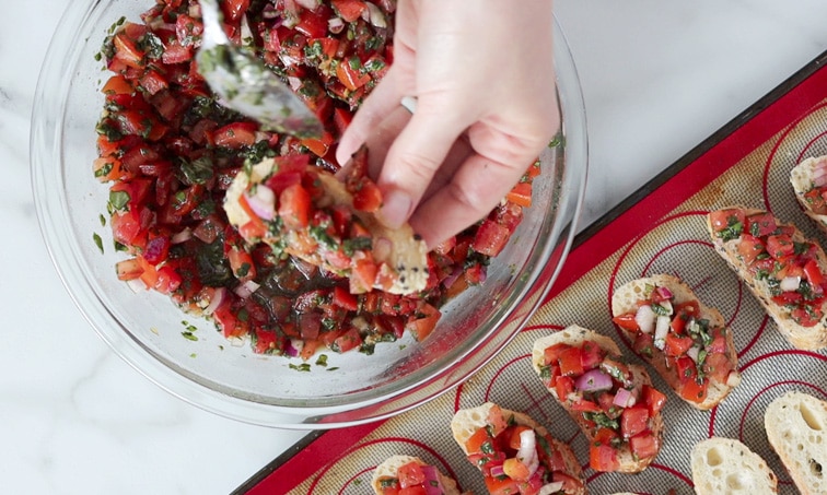 Hand holding a slice of baguette over a large bowl of tomato-basil bruschetta mix with a spoon loading the baguette slice with tomato mix. Bottom right corner of image shows a baking tray with prepped baguette pieces in a row.