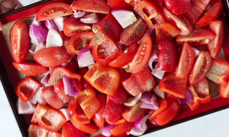 Prepped baking tray of tomatoes, red peppers and red onion, with seasoning sprinkled over top, ready for baking.