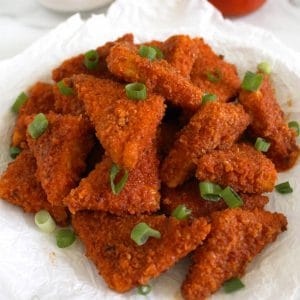 Close up of a pile of breaded, triangular tofu nuggets that are covered in a red sauce and topped with a sprinkle of chopped green onion.