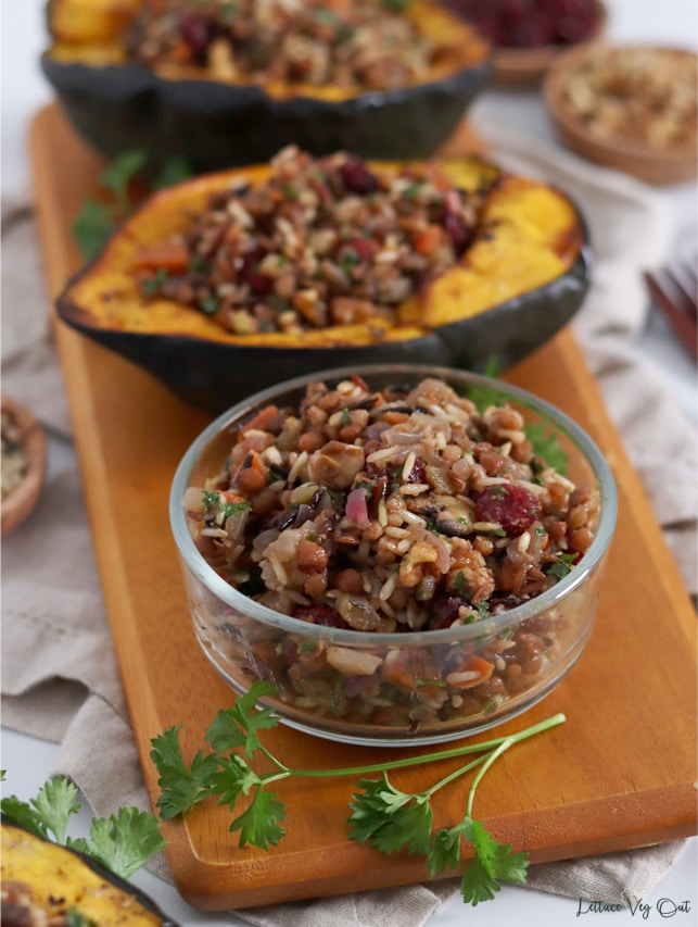 Small glass bowl filled with wild rice, cranberry and walnut stuffing, sitting on a long wood board with two squash halves filled with the stuffing.
