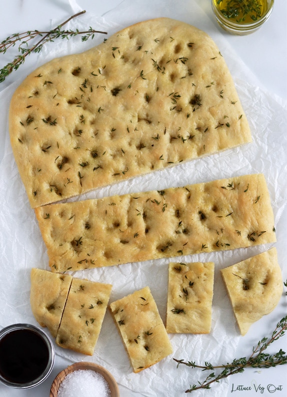 Top view of a loaf of focaccia bread on crumpled parchment paper. One row is cut into squares, another row is cut whole and half the loaf is uncut. Fresh thyme and jars of olive oil, salt and balsamic decorate around the bread.