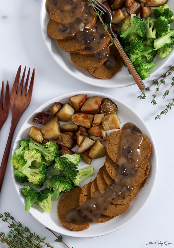 Top view of one full plate and one half-cropped plate each with half the plate covered with a row of sliced seitan with gravy on it and the other half of the plate covered with roast potatoes and broccoli. Wood forks and fresh thyme decorate image.