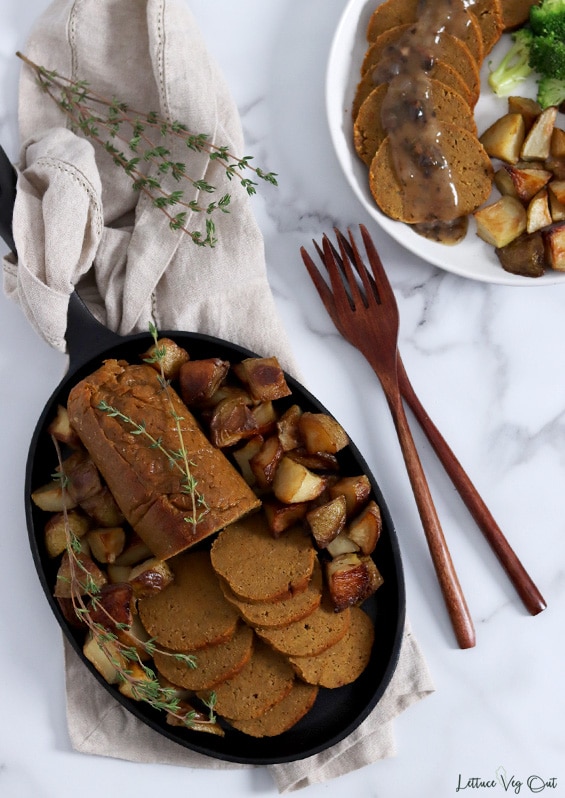Top view of an oval cast iron pan filled with a log of roast seitan that is half slices and surrounded by roast potatoes with thyme garnish. Top right corner of image shows a plate with a row of seitan topped with gravy next to roast potato and broccoli. Two large wood forks between the cast iron pan and the plate.