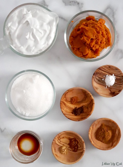 Top view of an arrangement of ingredients in glass and wood dishes. From top left working right then down: coconut cream; pumpkin puree; granulated sugar; mace and allspice; cream of tartar; vanilla extract; ground ginger and nutmeg; cinnamon.