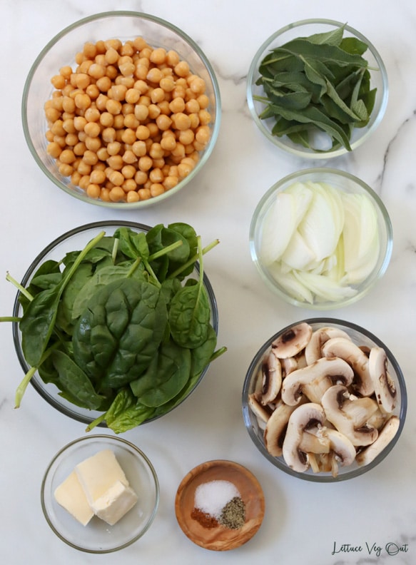 Top view of an arrangement of ingredients in glass dishes. From top moving left then down: chickpeas; sage; sliced onion; spinach; sliced mushrooms; vegan butter; salt + pepper + nutmeg.