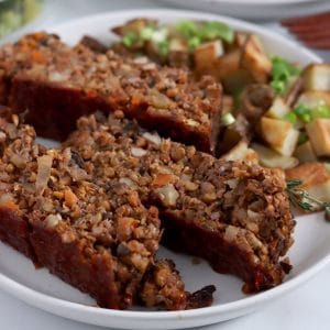 Square cropped image of three slices of lentil loaf that has a rough texture with flecks of lentils and vegetables throughout, sitting on a plate with roast potatoes topped with green onion sitting to the back of the plate.