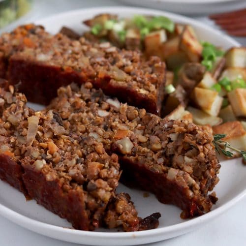Three slices of lentil loaf that has a rough texture with flecks of lentils and vegetables throughout, sitting on a plate with roast potatoes.