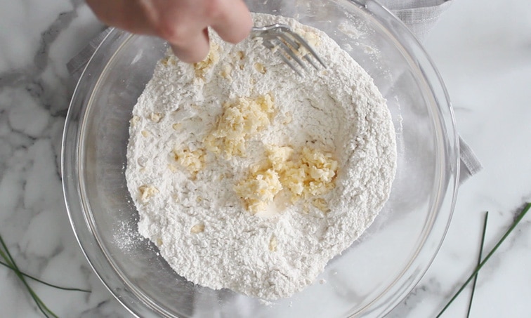 Top view of a large glass mixing bowl filled with flour and grated butter pieces. A hand holding a fork is mixing the butter into the flour.