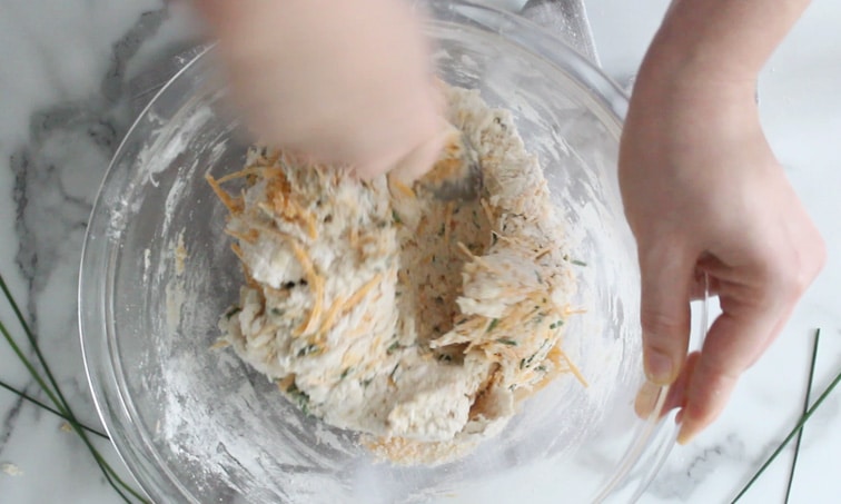 Top view of a large glass bowl filled with cheese and chive scone dough being mixed with a spoon.