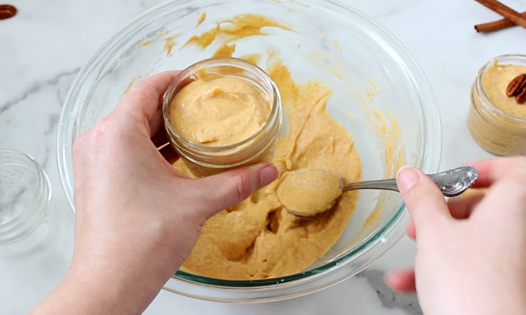 Scooping pumpkin mousse from a large bowl into a small jar that is mostly filled with mousse.