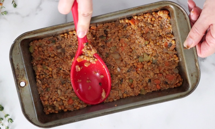 Top view of a loaf pan filled with a lentil and vegetable mixture that is being pressed into the pan with a large red spoon.