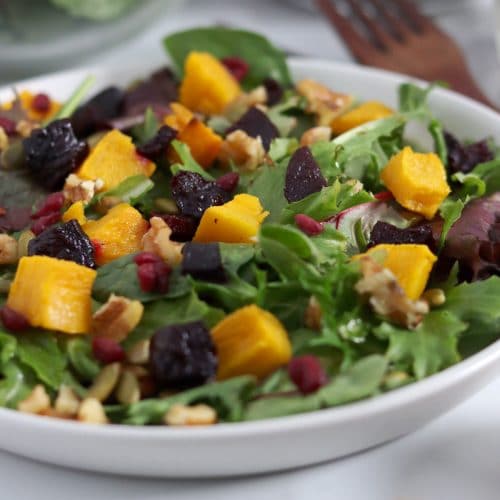 Square cropped image of a close up of a plate of salad topped with cubes of roasted pumpkin and roasted beetroot, pumpkin seeds, walnuts, pomegranate seeds and dressing.