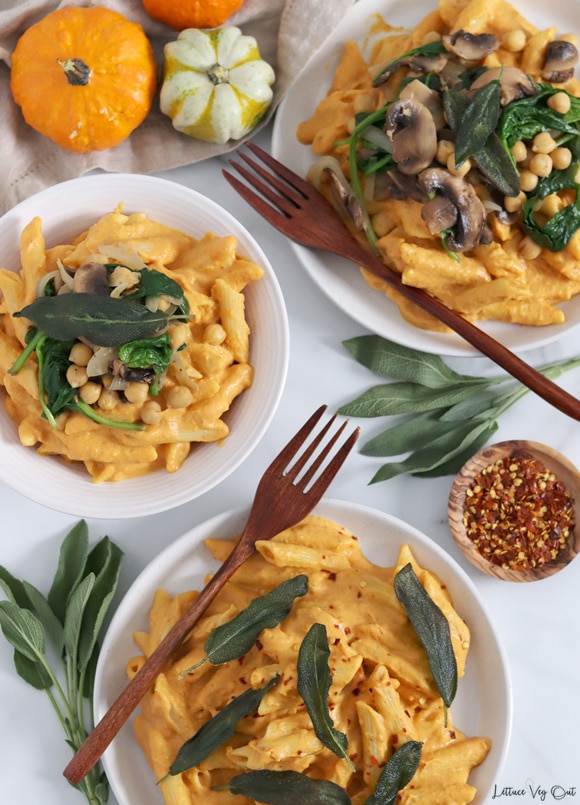 Top view of two plates and a bowl all filled with creamy pumpkin pasta sauce over penne noodles and garnished with fried sage leaves, sauteed spinach, mushrooms and chickpeas and wood forks on the two plates. Around the plates decorated with mini pumpkins, bunches of sage and a wood dish of red pepper flakes.