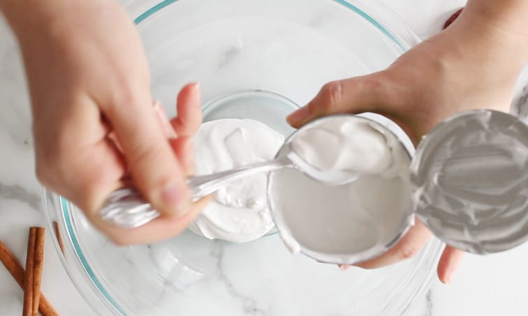 Top view of hands scooping coconut cream from a can into a large glass bowl below.