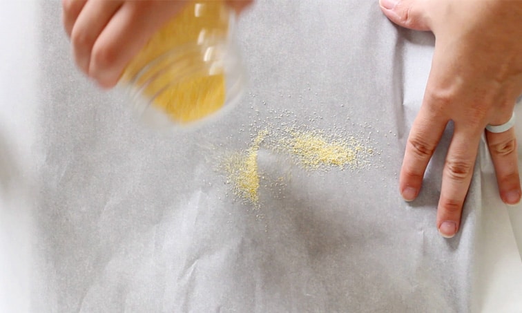Hand sprinkling cornmeal from a jar onto a piece of parchment paper while the other hand holds down the paper.