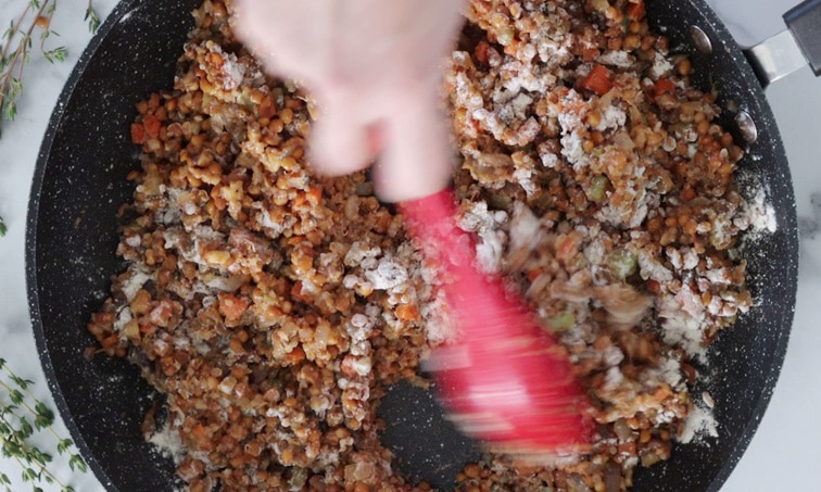 Top view of a pan filled with lentil and vegetable mixture with some flour sprinkled on top. A hand holding a large red spoon is stirring the flour into the lentil mix.