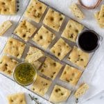 Square cropped image of the top view of a wire rack covered with square pieces of focaccia bread, a small jar of olive oil and small jar of balsamic vinegar.