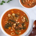 Square cropped image of a close up of a large bowl of minestrone soup (tomato broth, shell pasta, spinach, white beans, carrots and green beans). A second smaller bowl of soup is mostly cropped out at the top right of image.