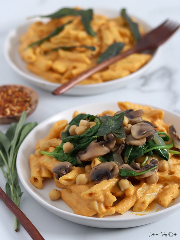 Close up of a plate of penne pasta coated with creamy pumpkin sauce and topped with sauteed onion, mushrooms, spinach and chickpeas. A second plate of pasta sits blurred in the back.