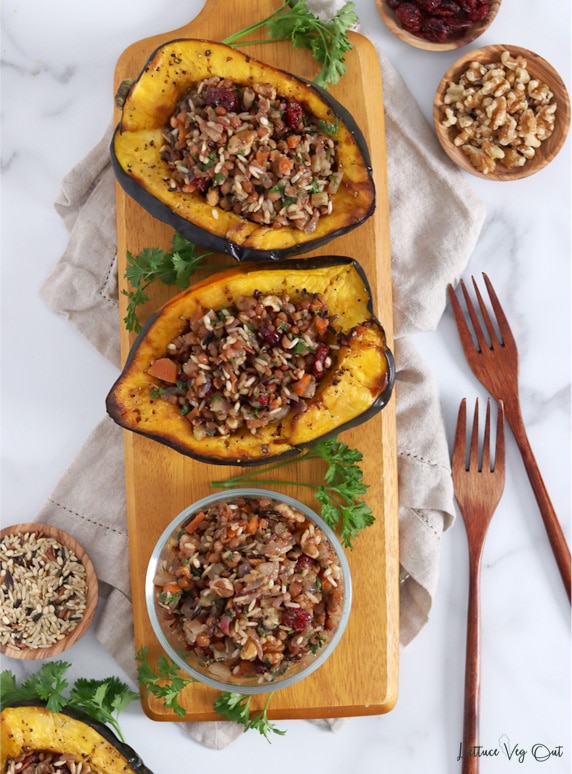Top view of a long wood board with two roasted squash halves stuffed with wild rice filling at the top and a glass dish of the stuffing at the bottom. Image decorated with fresh parsley, wood forks and small wood dishes with wild rice, cranberry and walnuts.
