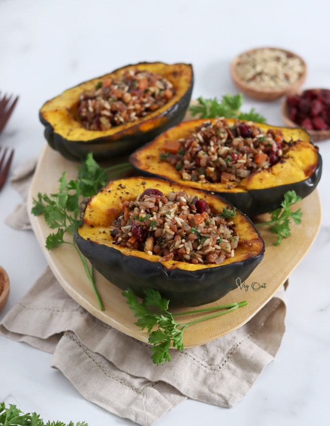 Three roasted acorn squash halves that are stuffed with wild rice, cranberry and walnut filling sitting on an oval wood board that is garnished with parsley. Board sits on a folded light brown towel.