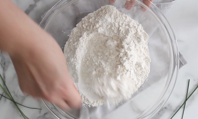 Top view of a hand mixing flour and other dry ingredients in a large glass mixing bowl.
