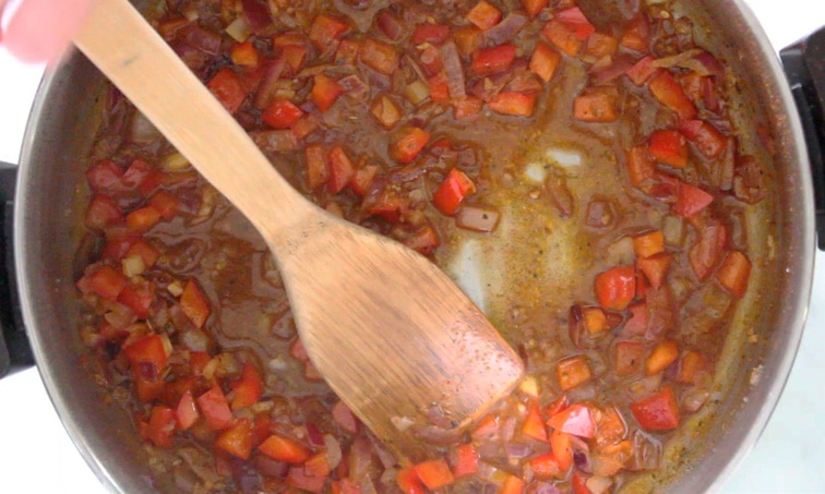 Top view of a pot filled with cooked red bell pepper and red onion coated in spices and a small amount of liquid. Hand stirs the pot with a wood spoon.
